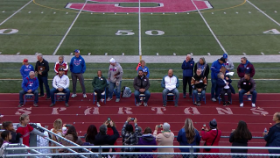 Inductees sitting on chairs in front of a crowd at a football game